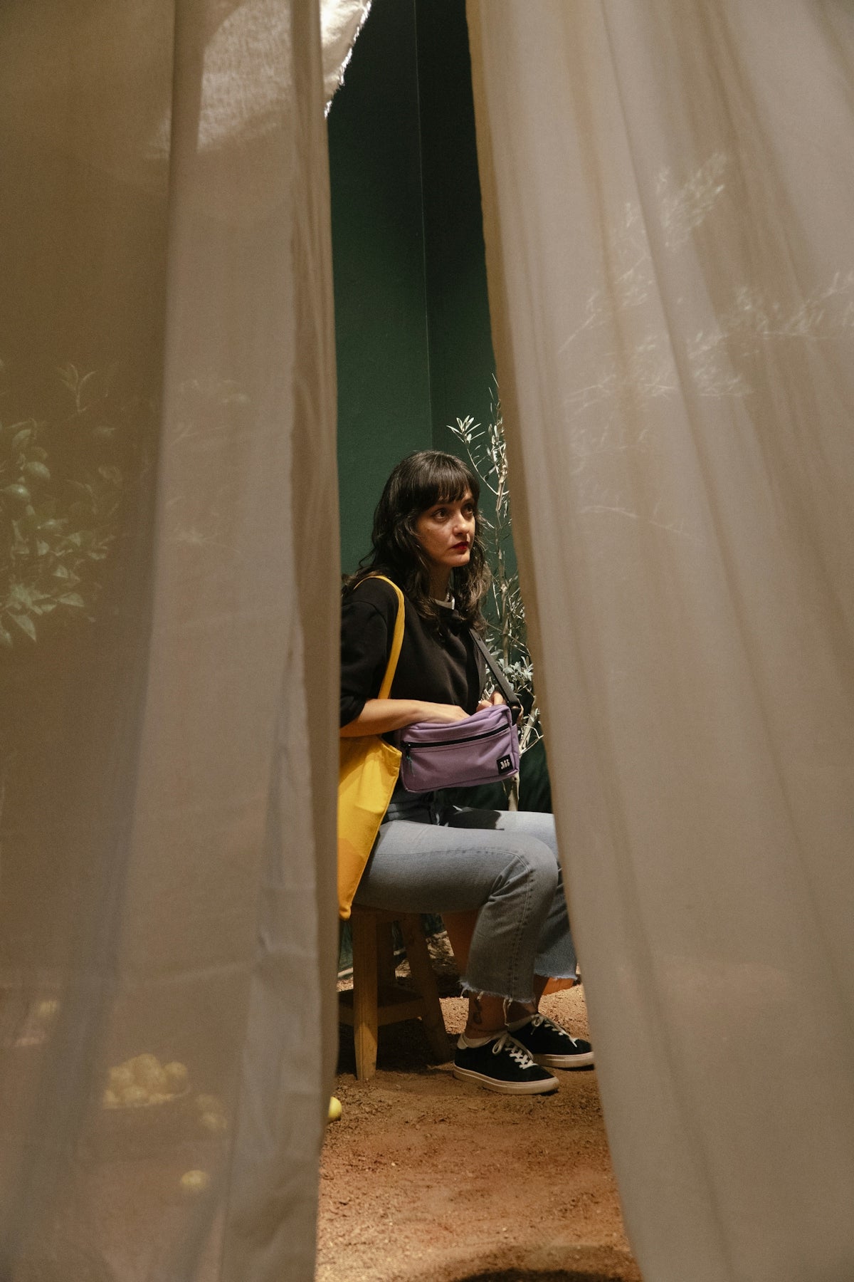 Woman sitting behind sheer curtains with a bag.