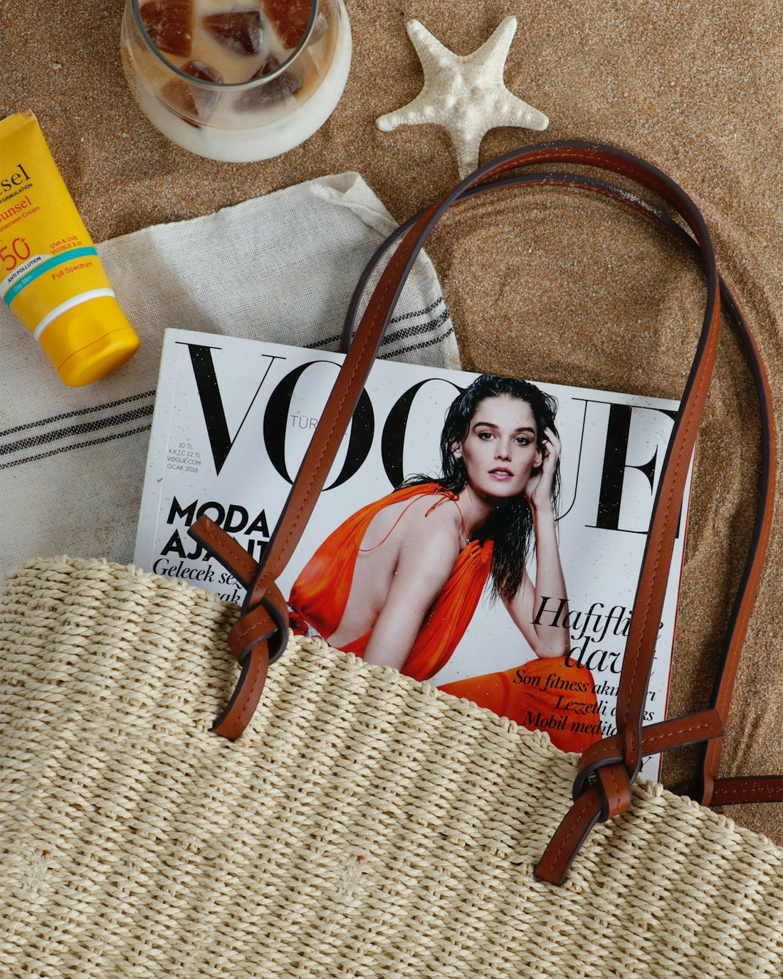 A straw tote bag, a fashion magazine, sunscreen, and a starfish on sand